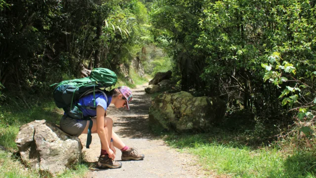 Ragazza seduta su una roccia con zaino in spalla che si sta allacciando una scarpa