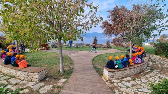 Parco Ecolandia con bambini seduti sulle panchine e adulti in piedi; prato verde e alberi tutt’intorno, cielo azzurro con nuvole leggere e il mare visibile all’orizzonte.