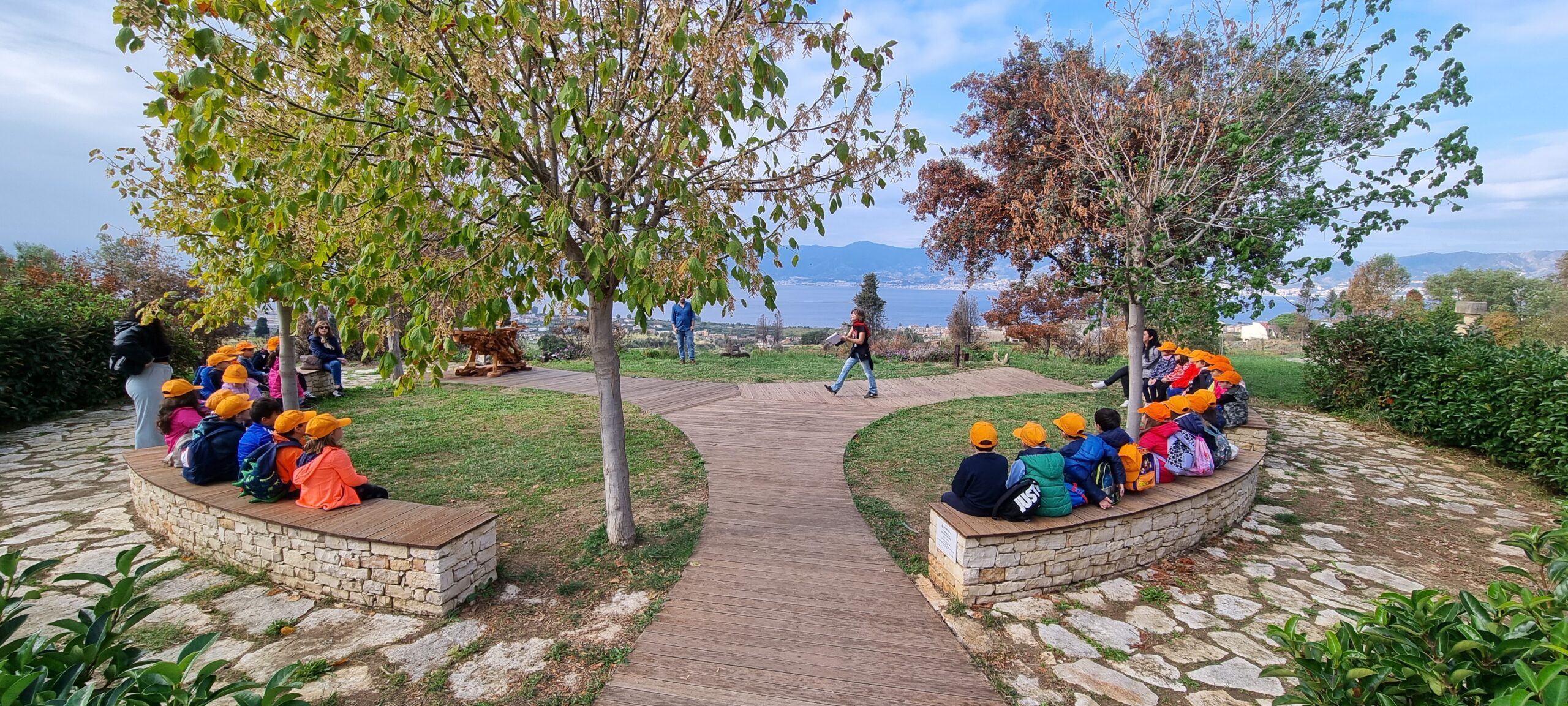 Parco Ecolandia con bambini seduti sulle panchine e adulti in piedi; prato verde e alberi tutt’intorno, cielo azzurro con nuvole leggere e il mare visibile all’orizzonte.