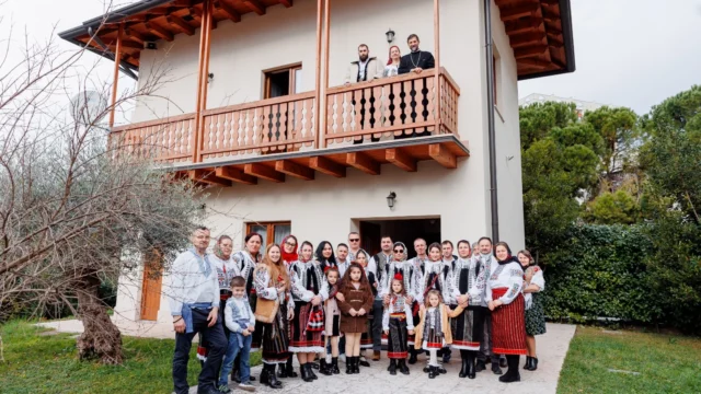 Gruppo di persone, tra adulti e bambini, in abiti tradizionali folkloristici, posa davanti allo studentato con balcone in legno mentre tre persone osservano dall’alto del balcone.