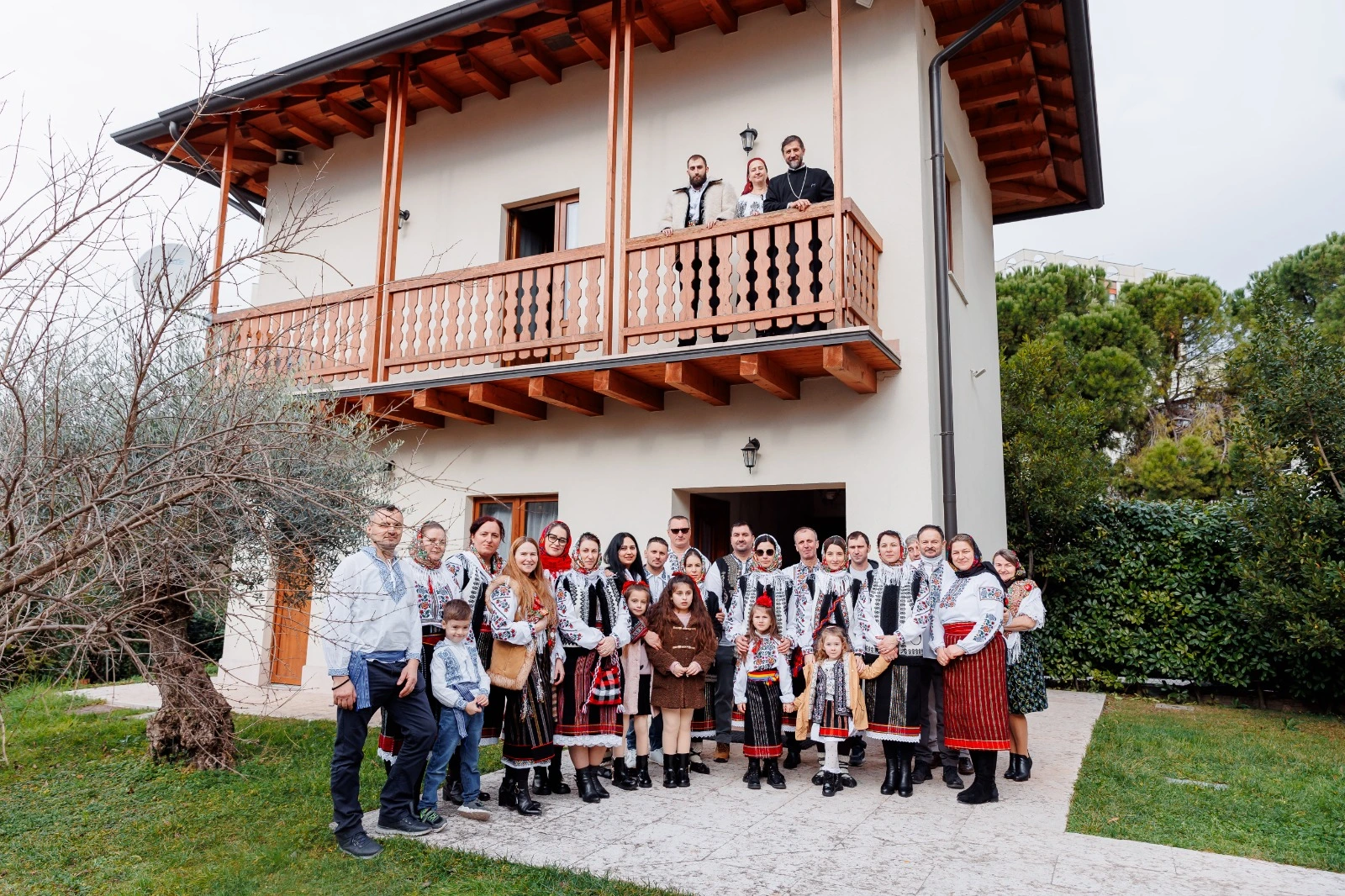 Gruppo di persone, tra adulti e bambini, in abiti tradizionali folkloristici, posa davanti allo studentato con balcone in legno mentre tre persone osservano dall’alto del balcone.