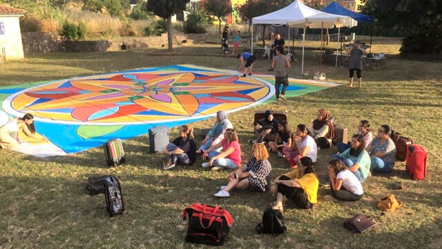 Gruppo di persone sedute sull’erba che osservano e partecipano alla realizzazione di un grande mandala colorato dipinto a terra, in un parco all’aperto con tende e alberi sullo sfondo.