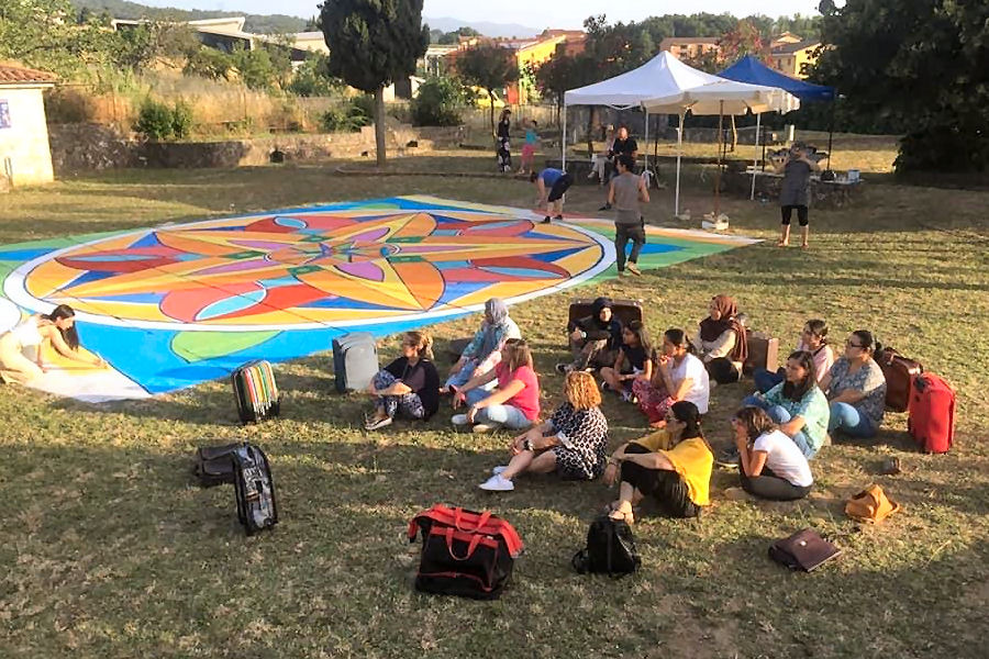 Gruppo di persone sedute sull’erba che osservano e partecipano alla realizzazione di un grande mandala colorato dipinto a terra, in un parco all’aperto con tende e alberi sullo sfondo.
