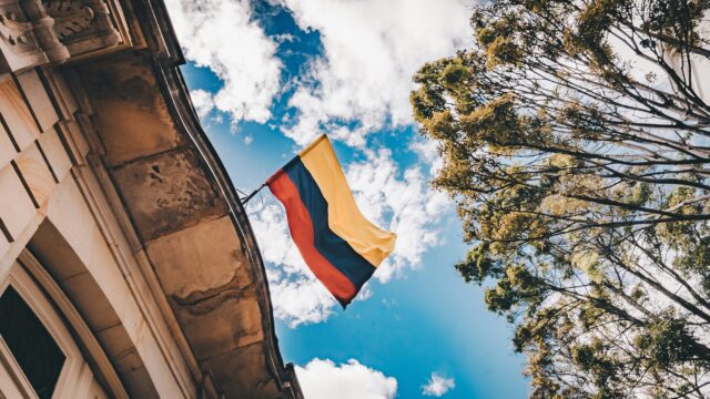 Bandiera della Colombia che sventola da un edificio in pietra, ripresa dal basso, con cielo azzurro e nuvole bianche tra alberi verdi.