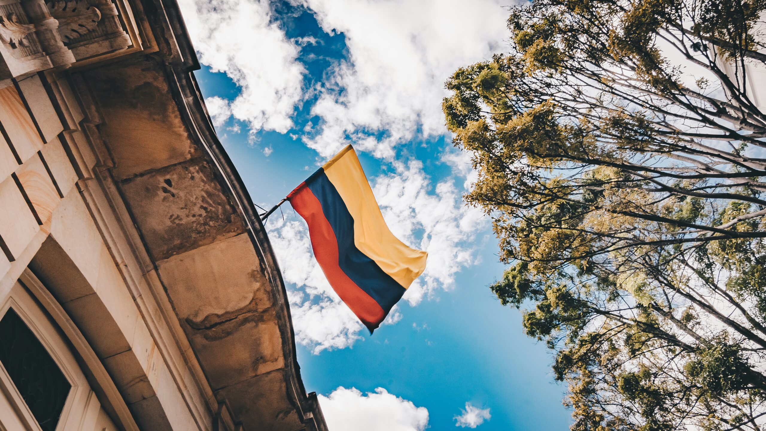 Bandiera della Colombia che sventola da un edificio in pietra, ripresa dal basso, con cielo azzurro e nuvole bianche tra alberi verdi.