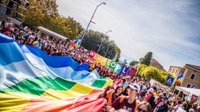 Corteo con grande bandiera arcobaleno e folla che marcia in strada.