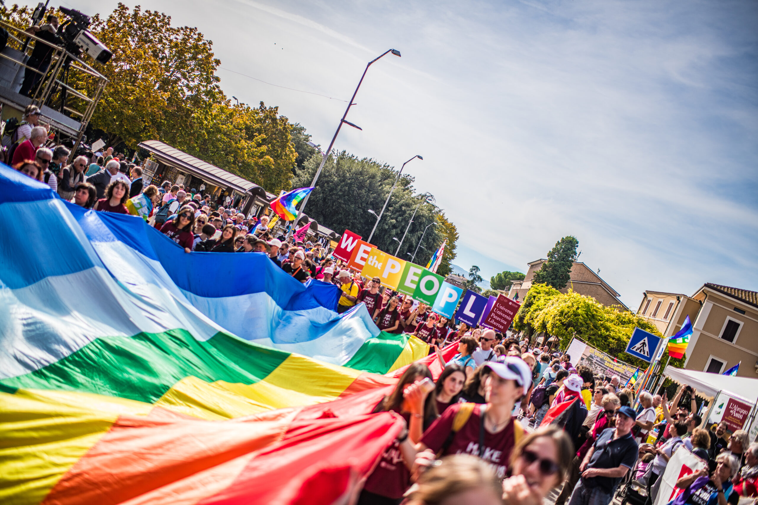 Corteo con grande bandiera arcobaleno e folla che marcia in strada.