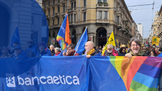 Corteo in piazza a Milano con partecipanti che reggono uno striscione di Banca Etica e bandiere.