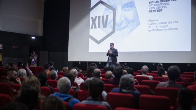 Pubblico in sala durante una presentazione del Sicilia Queer filmfest a Palermo, con relatore sul palco e schermo del festival sullo sfondo.