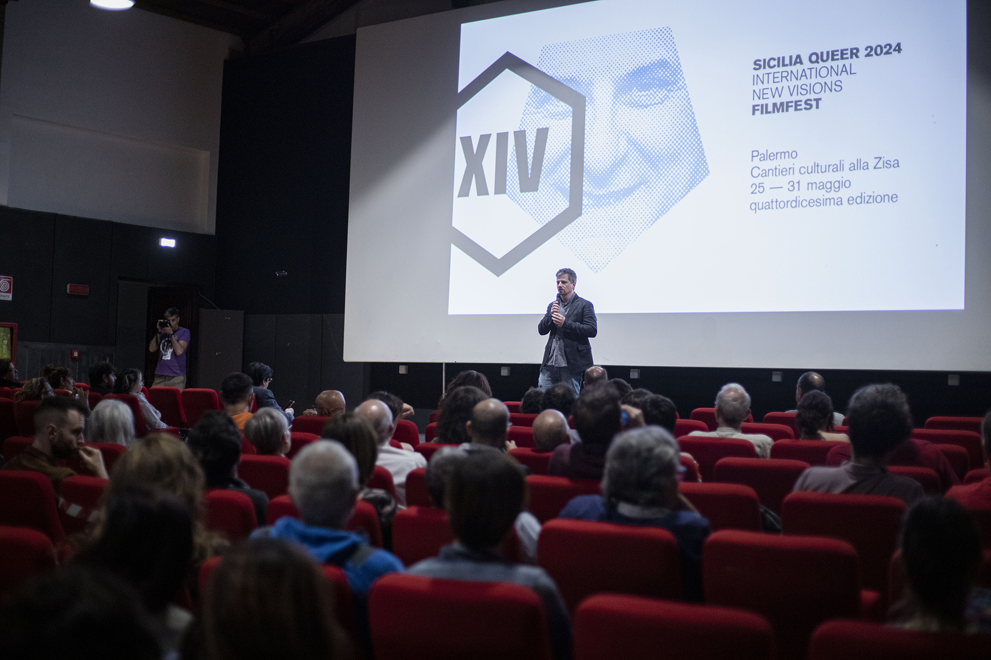 Pubblico in sala durante una presentazione del Sicilia Queer filmfest a Palermo, con relatore sul palco e schermo del festival sullo sfondo.