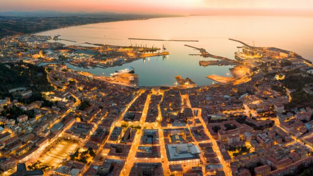 Panoramica aerea di città portuale italiana al tramonto, con centro storico illuminato e porto industriale affacciato sul mare.