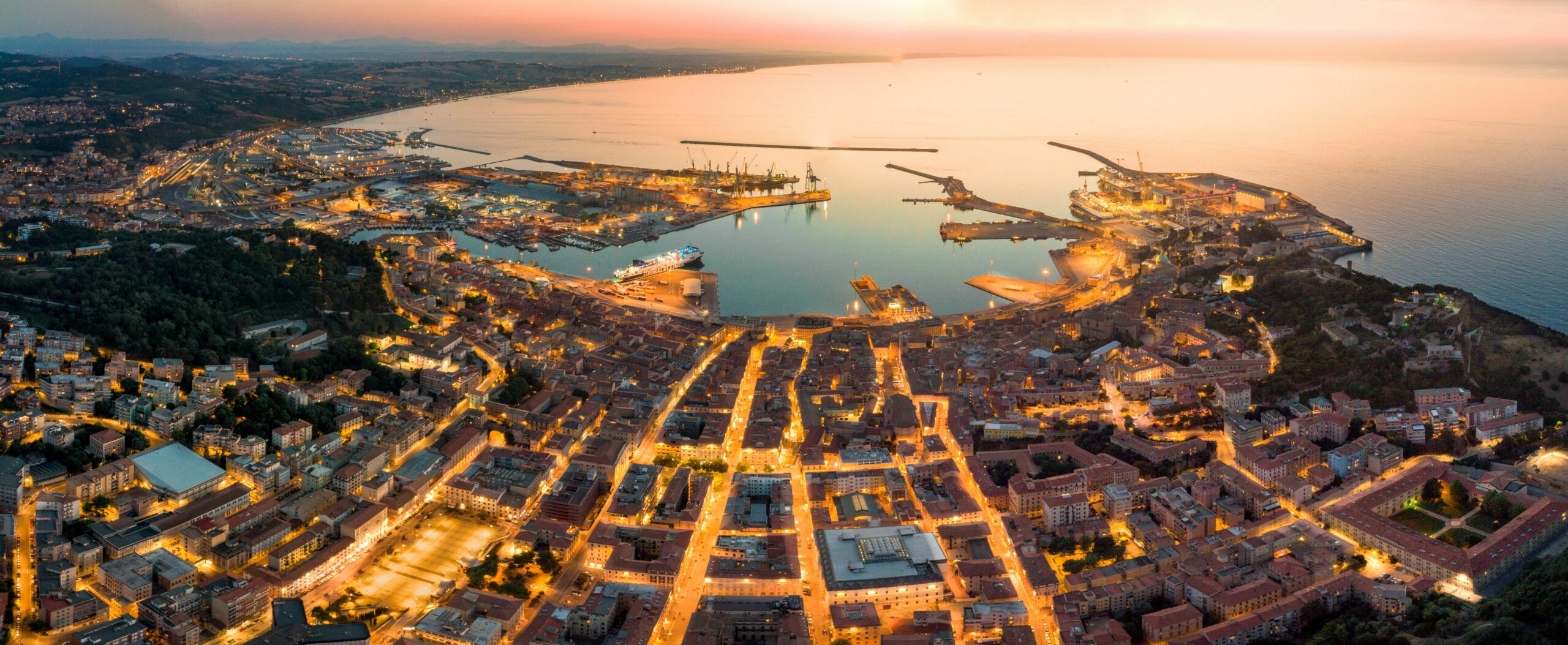 Panoramica aerea di città portuale italiana al tramonto, con centro storico illuminato e porto industriale affacciato sul mare.
