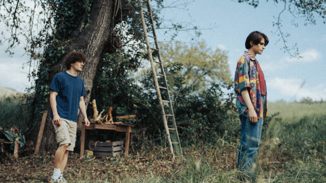 Scena film "La tana": Due ragazzi giovani si trovano in uno spazio verde all'aperto, vicino a un grande albero coperto di edera.Due ragazzi giovani si trovano in uno spazio verde all'aperto, vicino a un grande albero coperto di edera.
