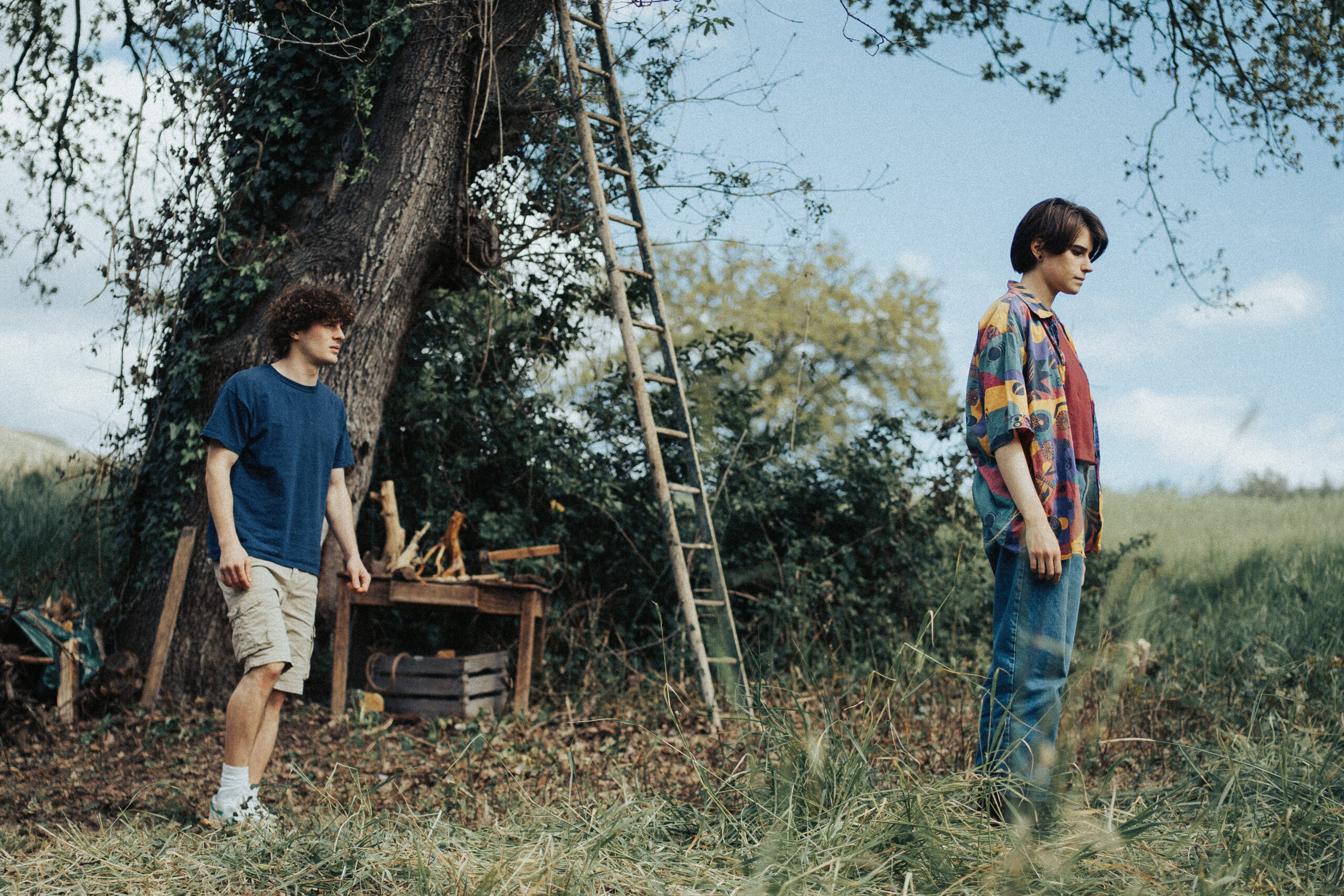 Scena film "La tana": Due ragazzi giovani si trovano in uno spazio verde all'aperto, vicino a un grande albero coperto di edera.Due ragazzi giovani si trovano in uno spazio verde all'aperto, vicino a un grande albero coperto di edera.