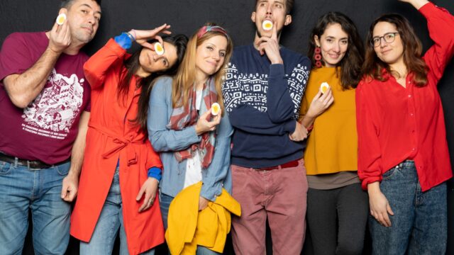 Un gruppo di sei persone posa davanti a uno sfondo scuro in modo giocoso, ciascuna tenendo o mostrando una metà di uovo sodo.